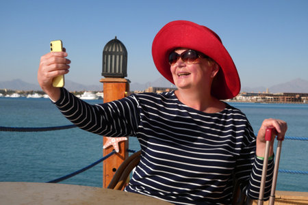 Senior woman in red hat and sunglasses waiting your a cruise ship in marina and making selfie by mobile phone, adventure is less, travel for pensioners, copyspace, outdoorsの写真素材