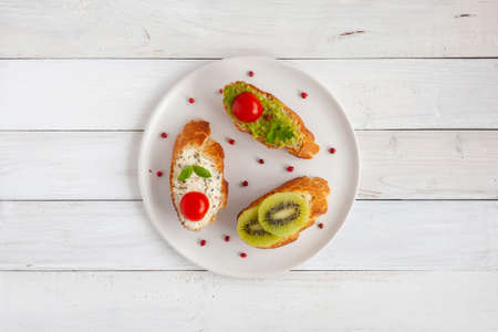 three homemade bright mini sandwichs with cream cheese and vegetables, on a white plate on wooden background, top viewの写真素材