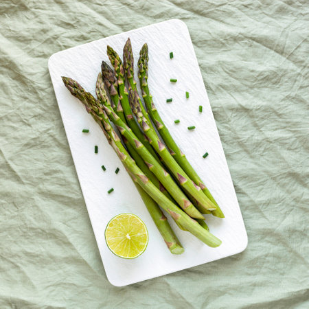 asparagus stems on the white rectangular plate served with green lemon and cut chives, top view, green linen background, square photoの写真素材