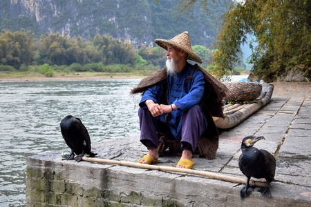 YANGSHUO, CHINA CIRCA APRIL 2017: The man with the straw hat and two cormorants sit on the bank of the river.のeditorial素材