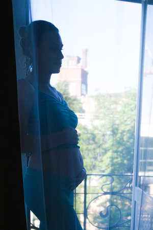 Attractive beautiful pregnant girl stands at the window behind the curtainの写真素材