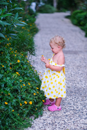 A young girl in a yellow polka dot dress is standing in a garden, looking at a flowerの写真素材