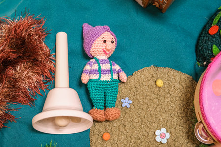 A small crocheted figure is sitting on the ground next to a bell. The scene is playful and whimsical, with the crochet figure resembling a character from a children's storyの写真素材