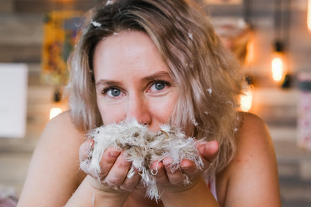 A woman is blowing feathers into her face. The feathers are white and fluffy. The woman has blonde hair and blue eyesの写真素材