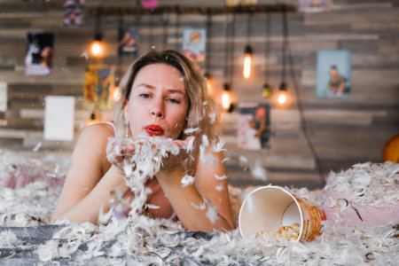 A woman is blowing feathers on a bed. The bed is covered in feathers and the woman is covered in themの写真素材