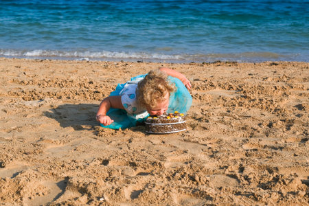 A young girl is eating a piece of cake on the beach. The cake is placed on the sand, and the girl is bending over to take a bite. The scene is lighthearted and playfulの写真素材