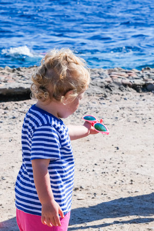 A young girl wearing a striped shirt and pink shorts is standing on a beach holding sunglasses. The scene is bright and sunny, with the ocean in the backgroundの写真素材
