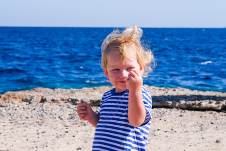 A young blonde child is standing on a beach, smiling and holding his hands up. The scene is bright and cheerful, with the ocean in the background and the child's hair blowing in the windの写真素材