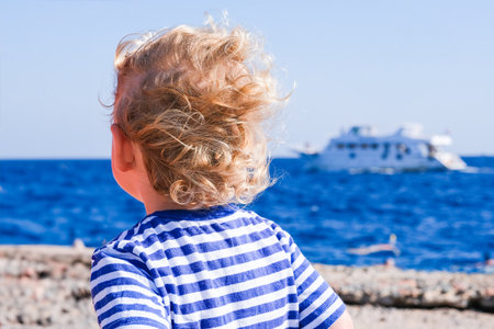 A child with blonde hair is standing on a beach looking out at a boat in the distance. The scene is peaceful and serene, with the child's hair blowing in the wind and the water in the backgroundの写真素材