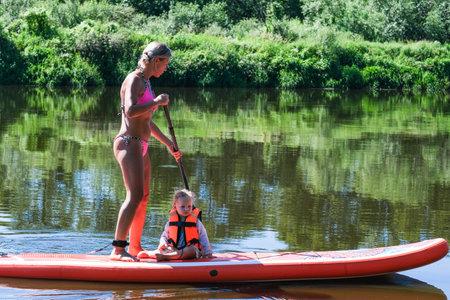 A woman and a child are enjoying a day on a paddleboard in a lake. The woman is holding the child on the paddleboard, ensuring their safety as they ride the waves. The scene is peaceful and sereneの写真素材
