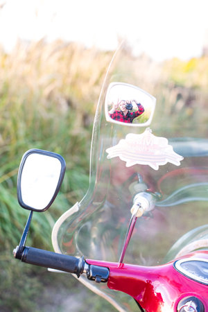 A man and woman are riding a motorcycle with a mirror on the handlebars. The mirror is reflecting the couple, and there is a sticker on the back of the mirror. The scene is set in a fieldの写真素材