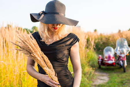 A woman wearing a black dress and a black hat is standing in a field of tall grass. She is holding a...