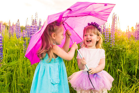 Two young girls are playing in a field with a pink umbrella. They are both smiling and laughing, enjoying their time together. The field is filled with purple flowers, creating a beautifulの写真素材