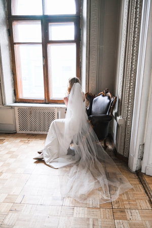 A woman wearing a white dress and a white veil sits in front of a window, looking out at the world. She is lost in thought, perhaps reflecting on her life or day. The chair in the room is blackの写真素材