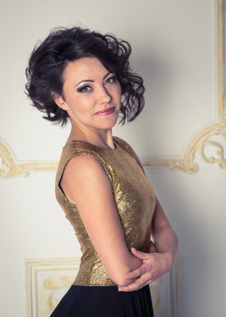 A woman in a gold dress is posing for a photo. She has a serious expression on her face and is standing in front of a white wall. Concept of elegance and sophisticationの写真素材