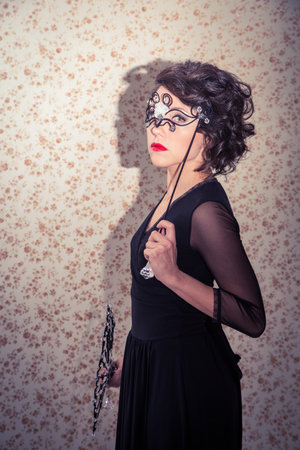 A woman in a black dress is holding a fan and wearing a mask. The image has a mysterious and elegant...