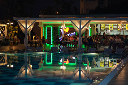 A pool with a neon sign that says "Time" above it. The pool is lit up and has a greenish tint. There are several people sitting at tables around the pool, and some are standingの写真素材