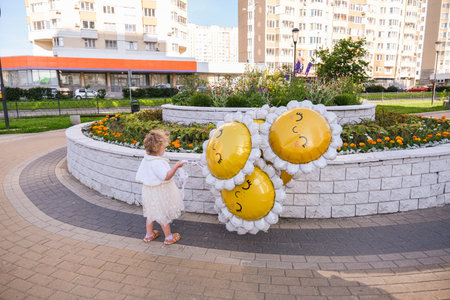 A little girl standing with daisy balloons by a flowerbed. Happy childhood concept and celebration moment. Outdoor leisure time.の写真素材