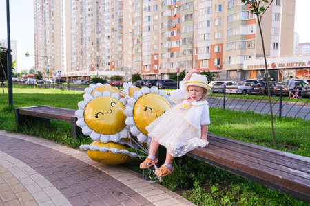 moscow, russia, 24.06.2025 Little girl in a hat and white dress sitting on a bench next to sunflower balloons outdoors. Children's birthday party.の写真素材