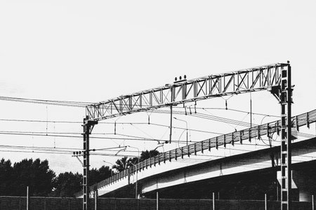 Moscow, Russia, 08.18.2025 Railway overhead power lines supported by metal structures and concrete bridge with trees in background. Monochrome urban transport infrastructure.の写真素材