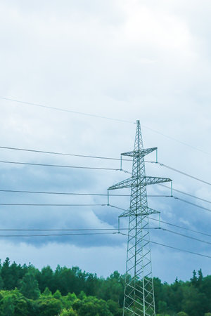 High voltage electric tower with power cables standing tall over lush green forest under a cloudy blue sky. Energy transmission concept.の写真素材
