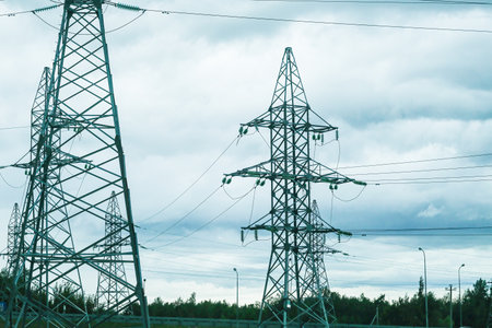Many electric power transmission line towers stand against a cloudy sky. Industrial high voltage...