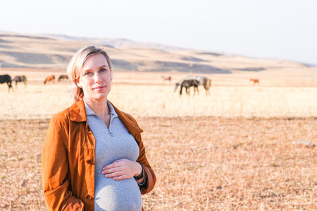 Caucasian pregnant woman touching her belly with horses grazing behind. Pregnancy journey in nature....