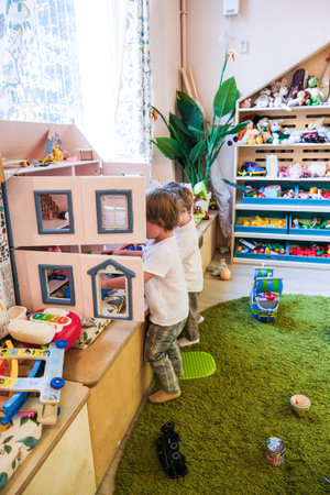 Two children are playing with a dollhouse in a room with lots of toys. The room is filled with various toys, including a teddy bear and a toy car. The children are standing on a rugの写真素材