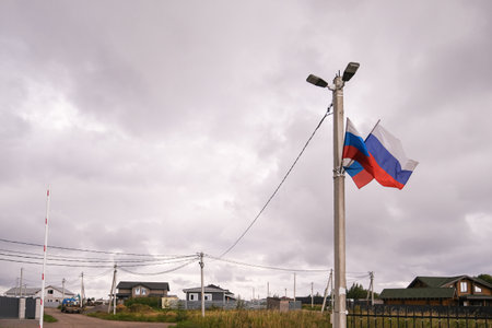 Russia, Saint Petersburg, 08/23/2025 Pair of Russian flags flying from light pole in a village under cloudy sky. Community representation and patriotism in local setting.の写真素材