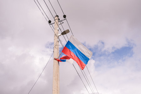 08/23/2025 Two flags with red, blue, and white stripes waving from an electrical pole with power lines and a street lamp against a cloudy sky.の写真素材