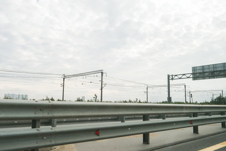 Road perspective of metal guardrail with overhead electrical wires and distant buildings under...
