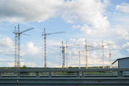 Row of construction cranes against a cloudy blue sky, representing development and urban growth,...