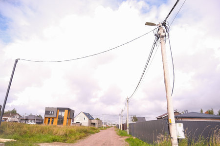 Russia, Saint Petersburg, 08/23/2025 Dirt country road leading past newly built house under construction and power line with tangled wires and street light on a cloudy day.の写真素材