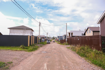 Russia, Saint Petersburg, 08/23/2025 Dirt road in a village with houses on both sides and utility poles stretching into the distance. Rural landscape for travel and homestead concept.の写真素材