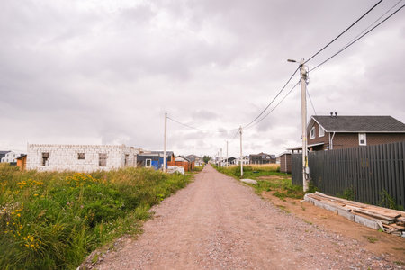 Russia, Saint Petersburg, 08/23/2025 Dirt road through a developing village with new houses and construction, showcasing rural development and housing progress under a cloudy sky.の写真素材