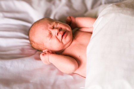 Newborn baby crying on white sheet. Tiny infant expressing discomfort and emotion during early development. Child care concept.の写真素材