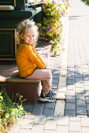 Blond hair little girl in a yellow sweater and sneakers sitting on a stoop. Happy child smiling on a sunny day outdoor. Childhood.の写真素材