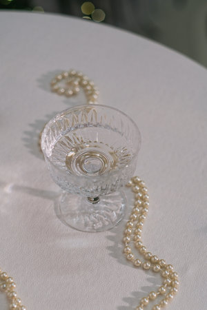 Elegant crystal glass with clear liquid and decorative pearl necklace on white tablecloth. Luxurious setting for celebration or special occasion.の写真素材