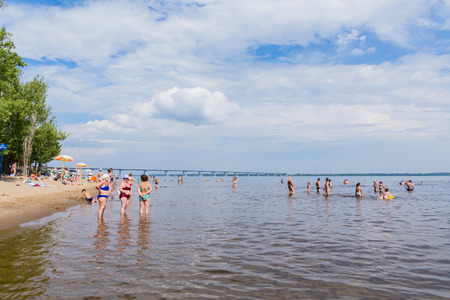 People sunbathe and swim on the sandy river beach. On a sunny summer day. White clouds on the blue sky. Picturesque landscape. Road bridge on the horizon.の写真素材