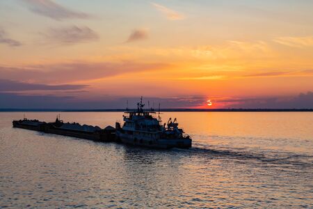 A tug on the river pushes a barge with sand or gravel. Sunset on the Volga river in Russia in July. Orange sun in the sky. Picturesque nature. Summer evening. River cruise.の写真素材