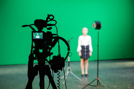 Filming in the studio on a green background for keying. A girl on stage. Video equipment: camera, tripod, microphone, spotlight, headphones.の写真素材