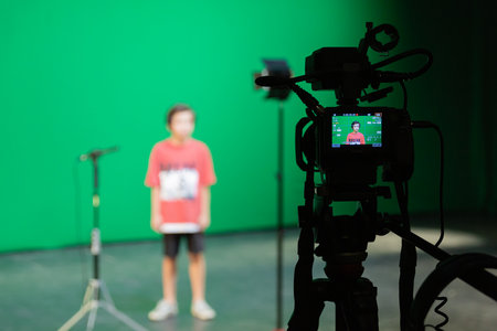 Video shooting in the studio on a green screen. A boy in a red T-shirt and shorts in the background. Shooting equipment: camcorder, microphone, tripod, spotlightの写真素材
