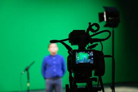 Videography of a boy in a blue shirt in the studio on a green background. Chromakey technology. A microphone on a stand, a video camera on a tripod and lighting equipmentの写真素材
