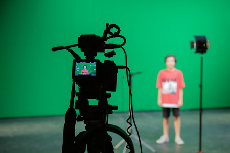 Video shooting in the studio on a green screen. A boy in a red T-shirt and shorts in the background. Shooting equipment: camcorder, microphone, tripod, spotlightの写真素材