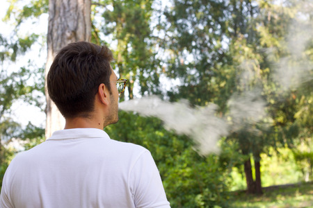young man emitting steam from the electronic cigarette, garden background defocusedの写真素材