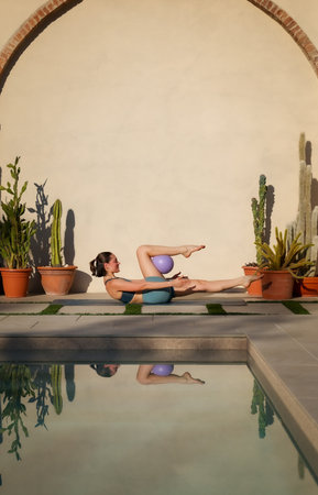 young pilates instructor doing exercise with lilac ball on mat near swimming pool, healthy lifestyle conceptの写真素材