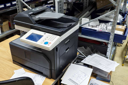 Close-up of a large printer with buttons on a table near a rack in a warehouse.