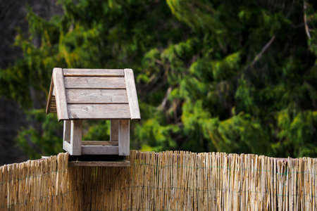Wooden bird feeder on a railing with spruce in the background and reeds on the railing.