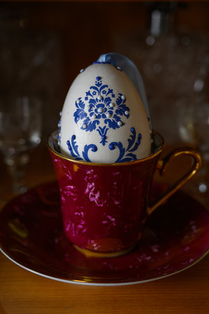 White goose Easter egg with blue pattern standing in a red cup and saucer with a golden handle.の写真素材