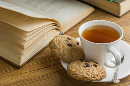 A cup of tea and some chocolate chip cookies over a books on a brown wooden table. Vintage style.の写真素材